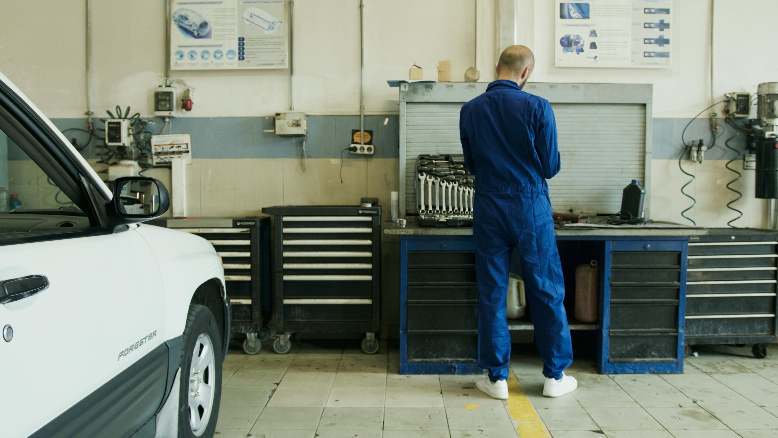 Mechanic in blue overalls inspecting a white car in a well-equipped garage.